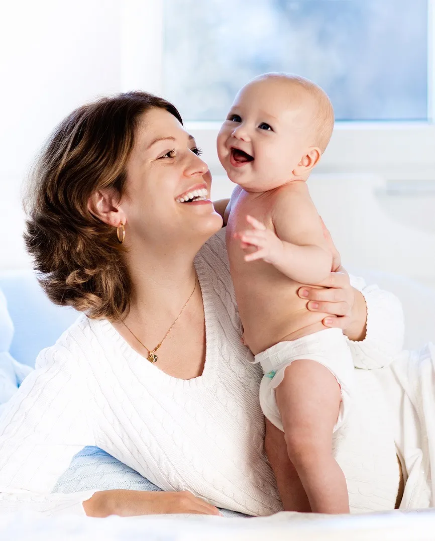 Smiling woman in white sweater holding and looking at a happy baby wearing a diaper indoors.