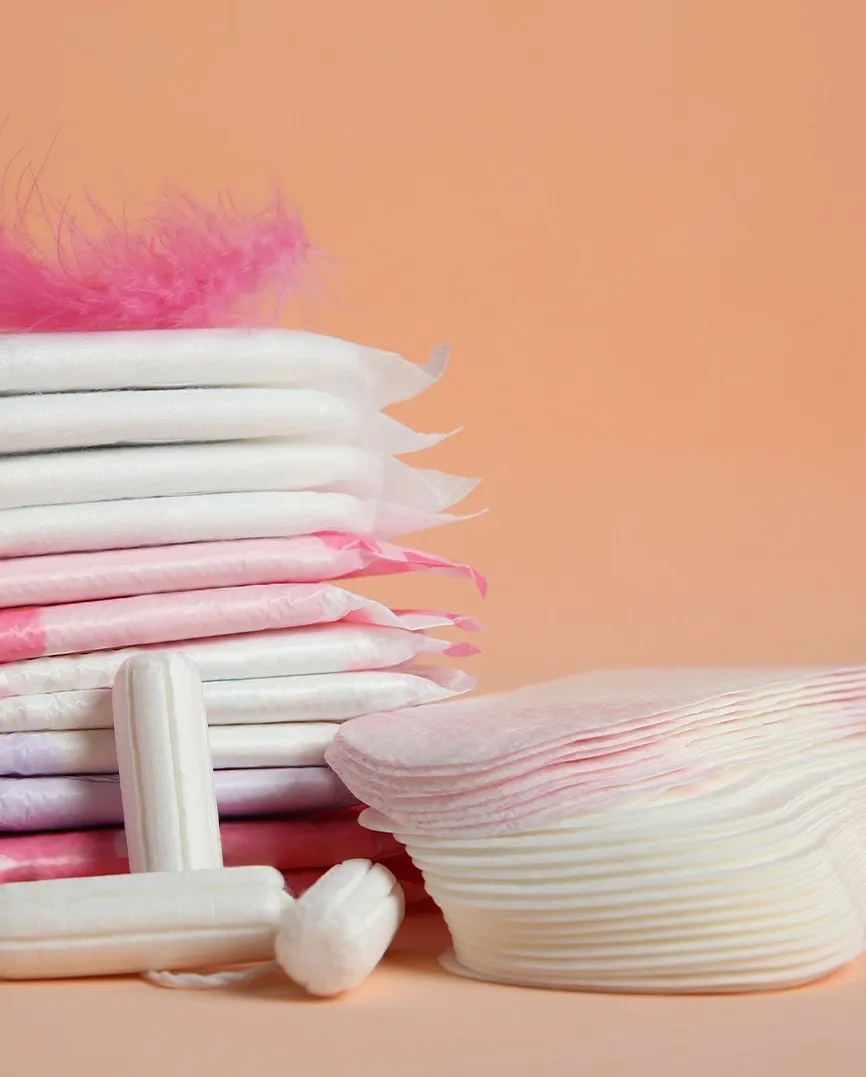 Stack of various feminine hygiene products including tampons and sanitary pads on a peach background.