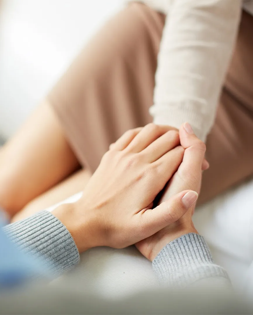 Two people holding hands for comfort, one wearing a gray sweater and the other in a brown outfit.