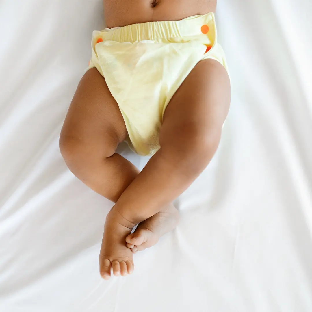 Infant wearing a yellow cloth diaper lying on a white surface with legs crossed.