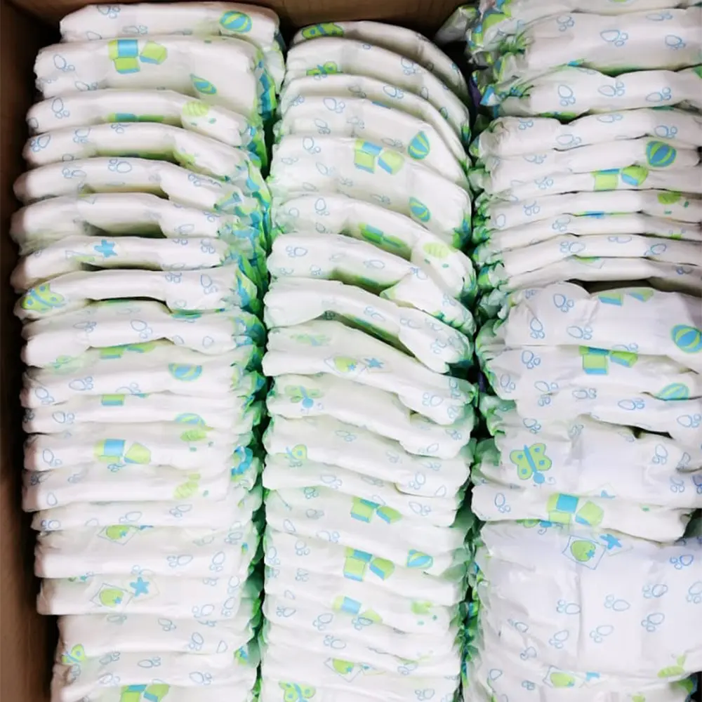 Stacked white baby diapers with blue and green playful prints inside a cardboard box.