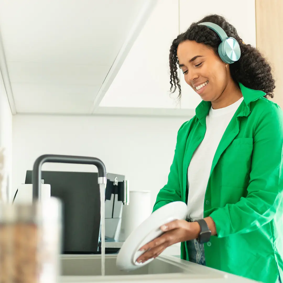 Smiling woman wearing green shirt and headphones washing a white plate at kitchen sink with running water.