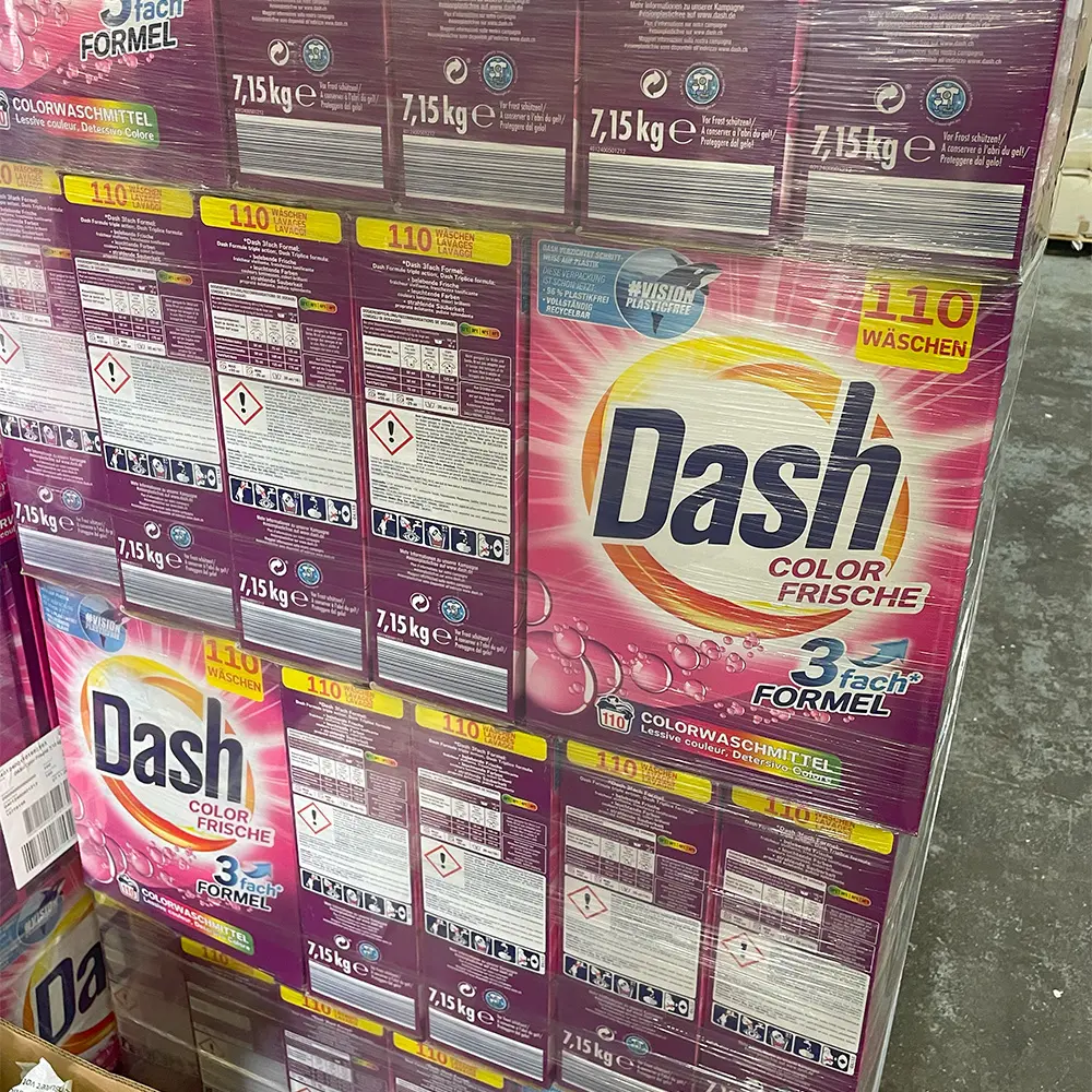 Stacked boxes of Dash Color Frische detergent with 110 washes and 3-fach Formel, packaged in purple and white wrap, displayed on a warehouse floor.