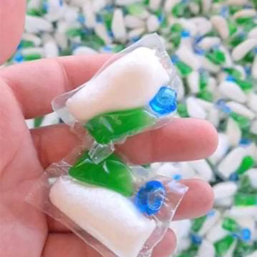 Hand holding two green, white, and blue laundry detergent pods with more pods scattered in the background.