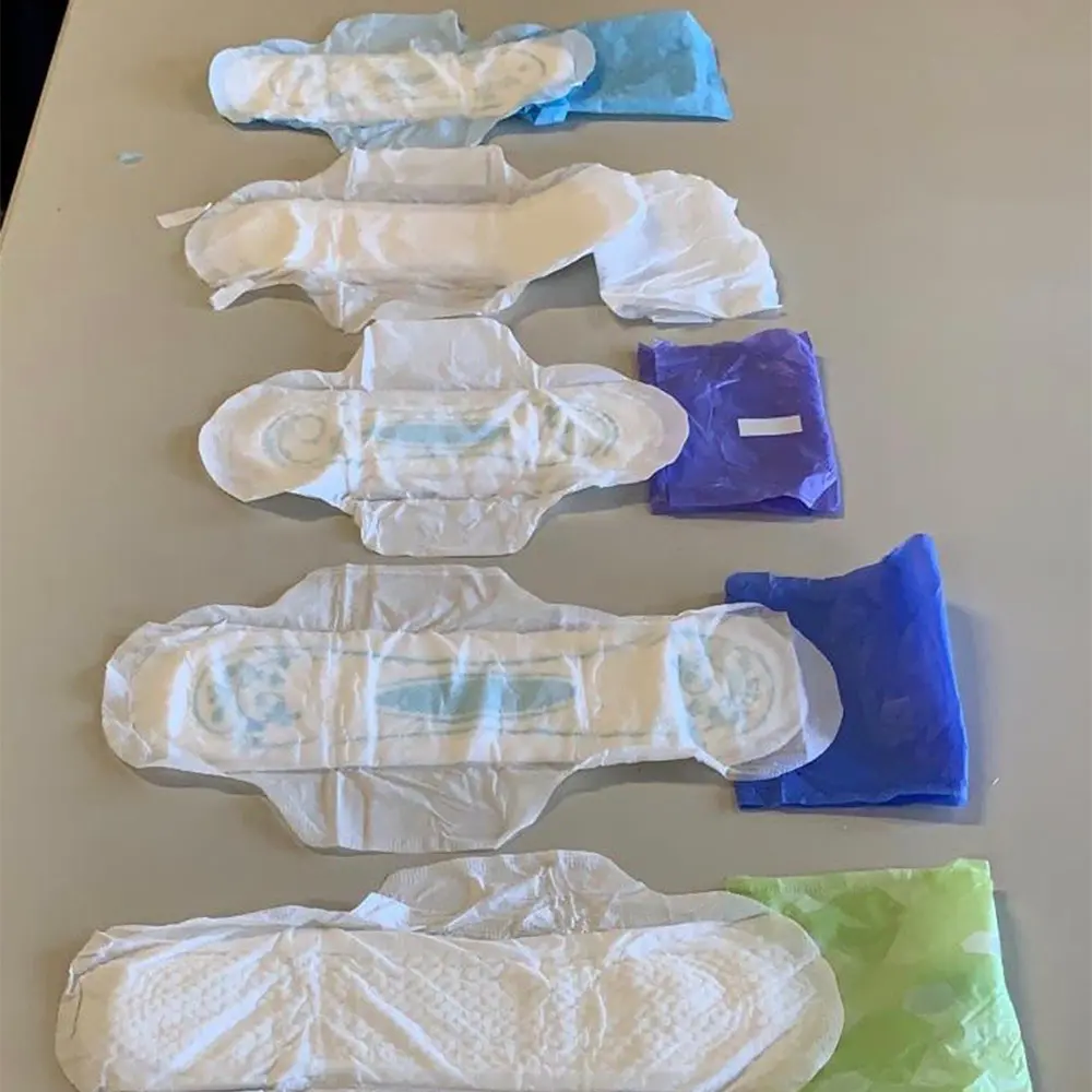 Five different sizes of unfolded sanitary pads arranged vertically on a table, each with its colored wrapper beside it.