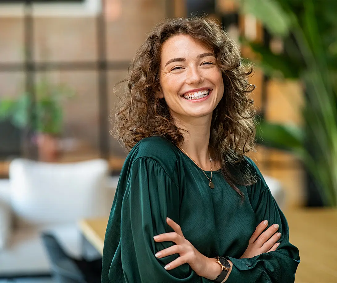 Smiling woman with curly hair wearing a dark green blouse and a watch, standing with arms crossed in a bright, modern indoor space.