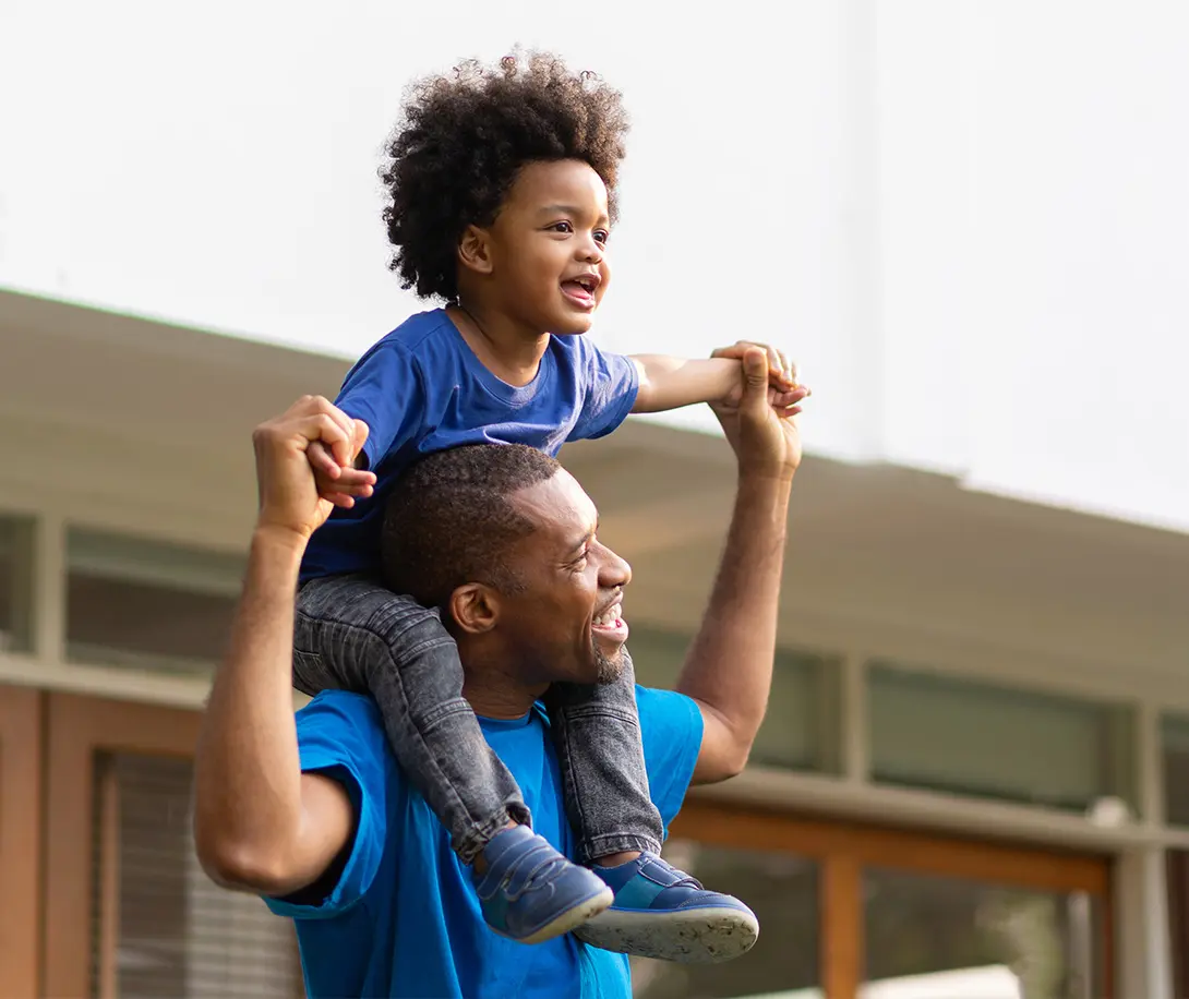 Smiling father carrying son on shoulders, holding his hands outdoors.