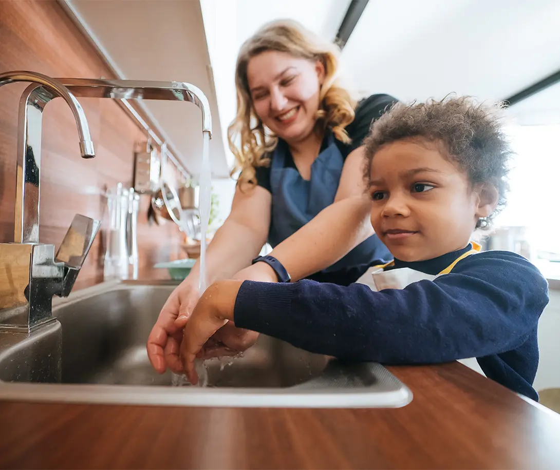 Woman smiling and helping young child wash hands under running kitchen faucet.