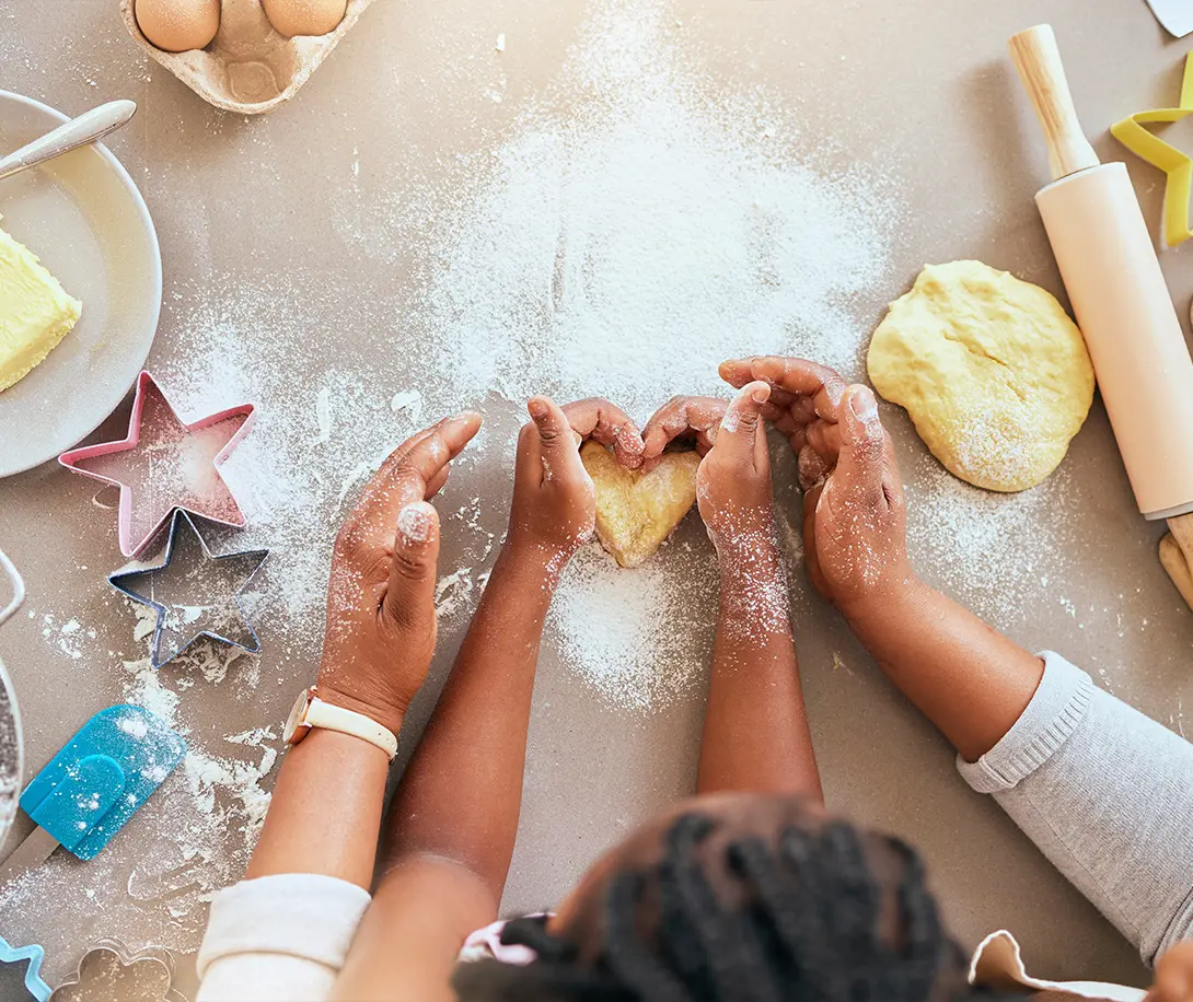 Adult and child hands shaping heart-shaped cookie dough on a flour-dusted surface surrounded by baking tools and ingredients.