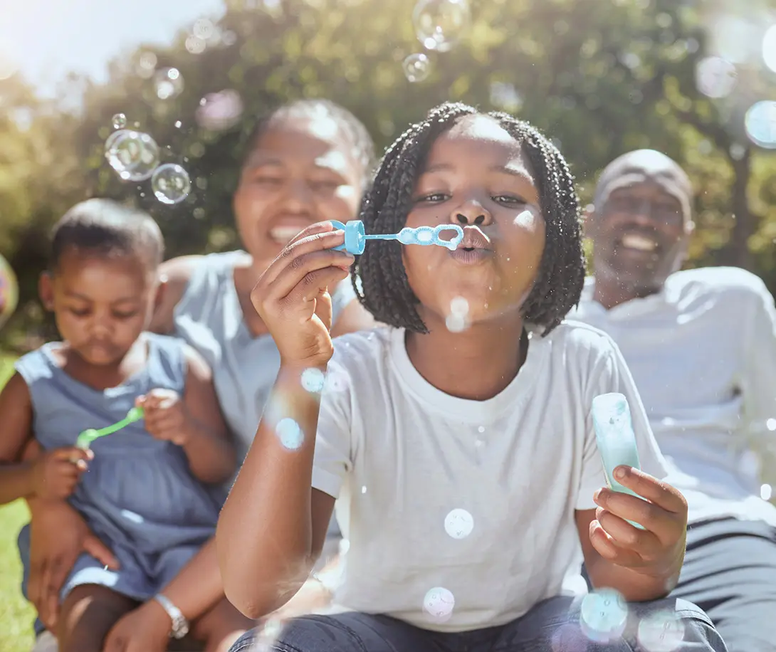 Child blowing soap bubbles outdoors with family sitting on grass in the background.