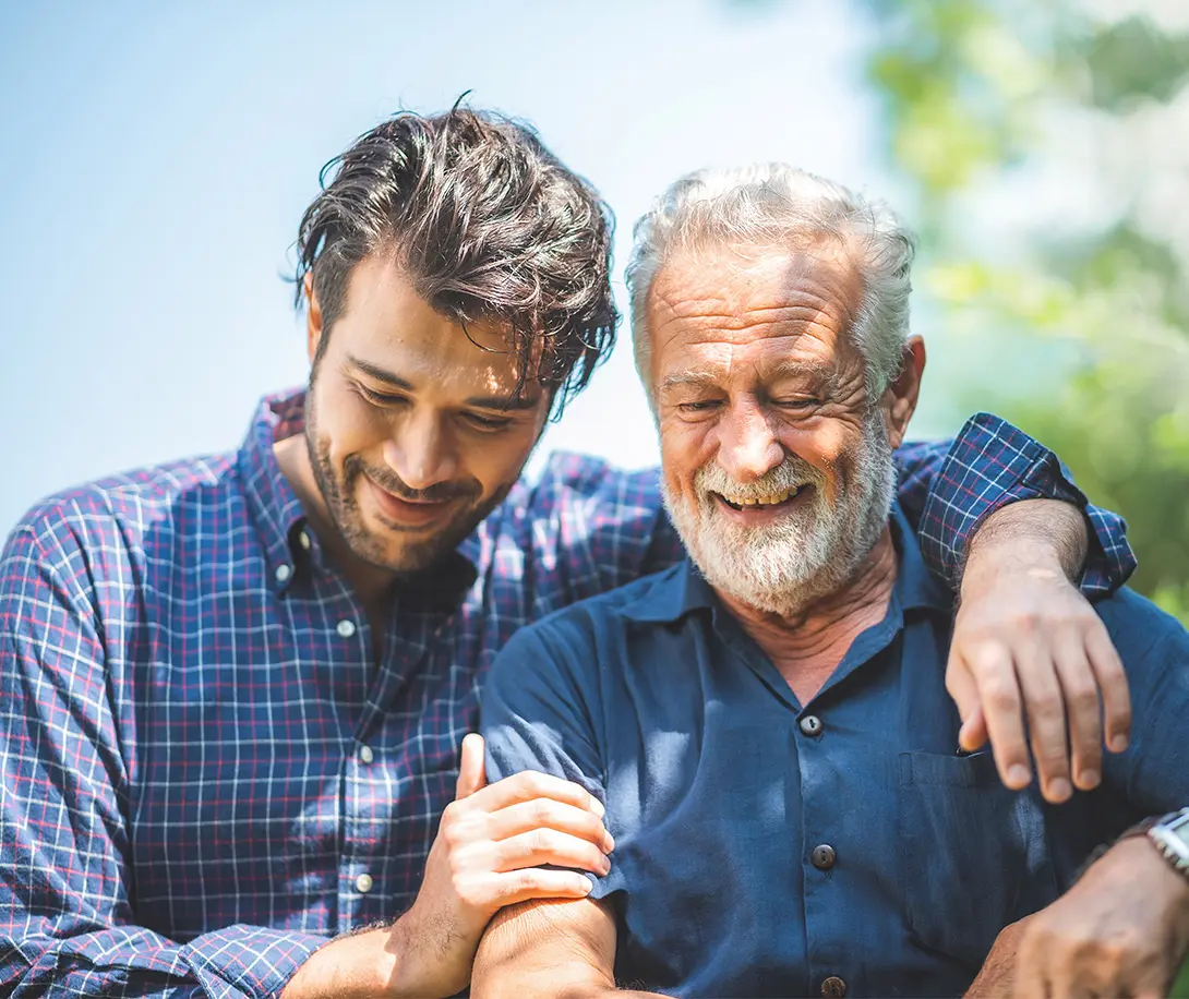 Smiling elderly man and younger man embracing outdoors with greenery in the background.