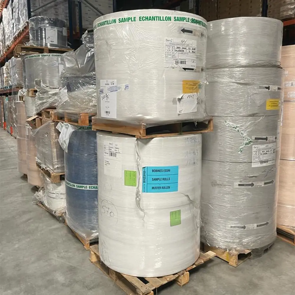 Large industrial rolls of material wrapped in plastic and stacked on wooden pallets inside a warehouse.