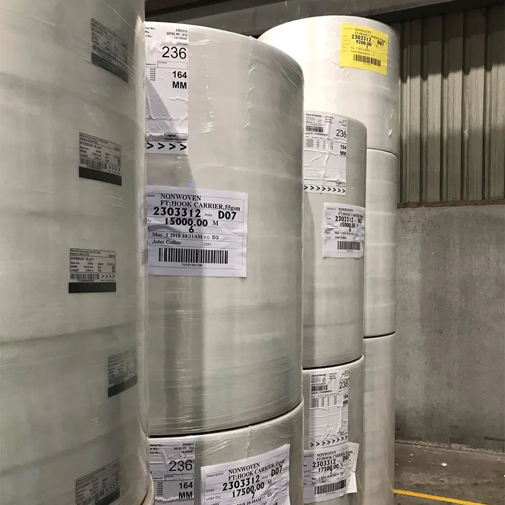 Large rolls of white nonwoven fabric stacked in an industrial warehouse with visible labels showing measurements and batch details.