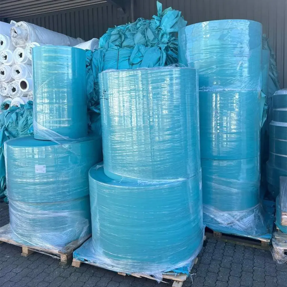 Large rolls of blue nonwoven fabric wrapped in plastic on wooden pallets in a warehouse.